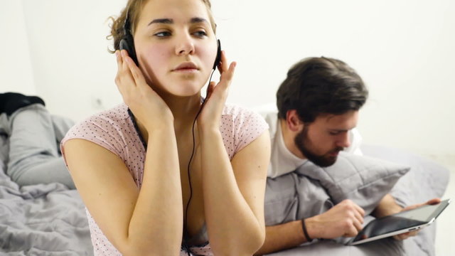 woman listening to music in headphones, man using tablet in bed