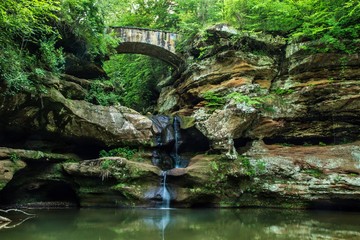 Fototapeta premium Enchanted Forest Waterfall. Stone bridge crosses over an enchanting forest waterfall. Hocking Hills State Park. Logan, Ohio.