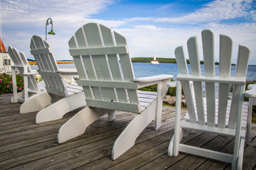 Island Life. Adirondack chairs line a wooden deck overlooking the Mackinaw Island waterfront with a lighthouse in the background. Mackinaw Island, Michigan.