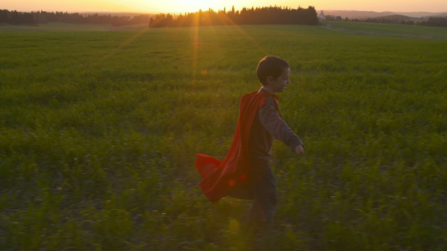 young boy with a superman cape running in a field during sunset