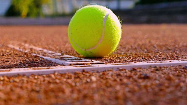 A rolling tennis ball stops at a line on a red tennis court