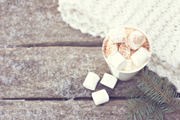 A cup of warm cocoa with marshmallows and a cozy knitted scarf on old wooden boards in the snow