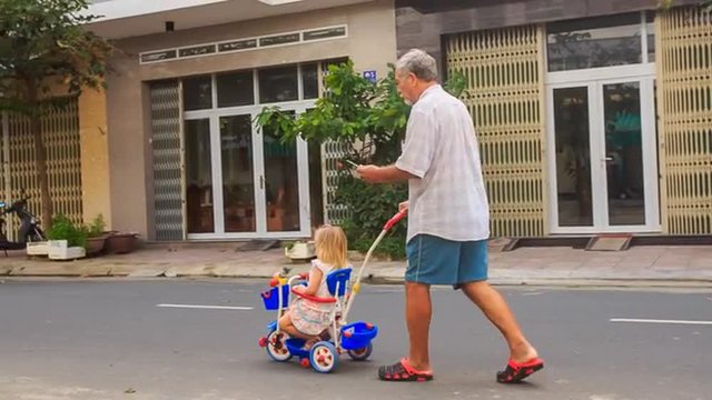 Grandpa Pushes Tricycle with Little Girl Reads Smartphone