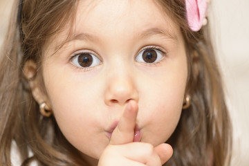 Portrait of expressive beautiful little girl finger up to lips for making a quiet gesture. Girl showing silence gesture looking at the camera