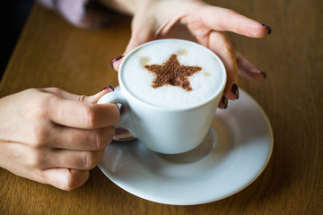 Women's hands with a hot cup of coffee. Coffee with milk, latte. A cup of coffee on the table. Embracing a cup of coffee. Cozy home atmosphere. Morning coffee. Toned image.