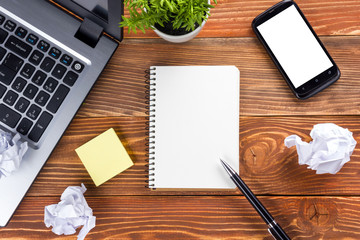 Office table desk with supplies, white blank note pad, cup, pen, pc, crumpled paper, flower on wooden background. Top view