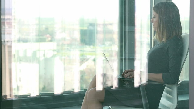 Businesswoman typing on laptop while sitting on chair behind glass wall