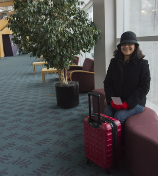 Hispanic young woman sitting on a bench in airport labby waiting for her flight to be called 

