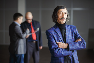 Old aged businessman wearing blue jacket standing with hands clasped. At background two men in...