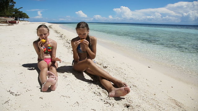 Two girl sisters sitting on a tropical beach,eat mango.children eats mango on the shore of tropical sea on a white beach.Travel concept.Family,summer vacation.Happy family