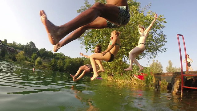 Four young friends running and jumping into river on beautiful sunny day, in slow motion