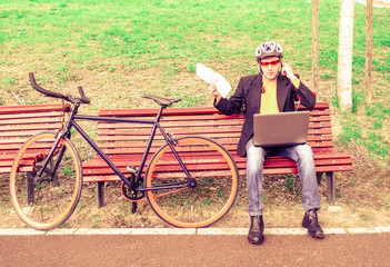 Business man working with laptop and using mobile phone with earphones sitting in a park bench next to sport bike - Young manager gesticulating outdoor - Concept of stress caused by work situations 