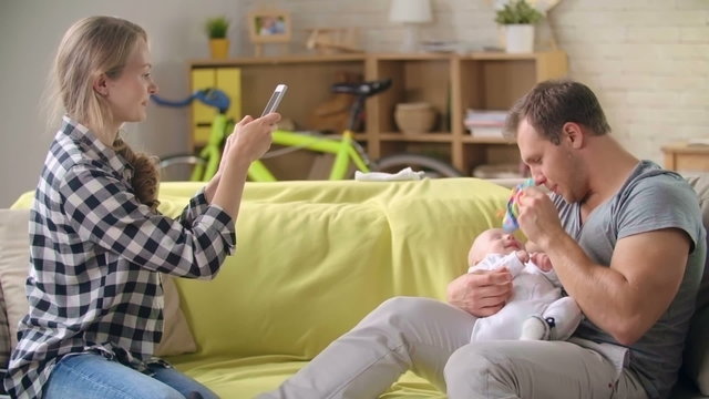 Young mother taking picture with smartphone of her husband playing with their little son