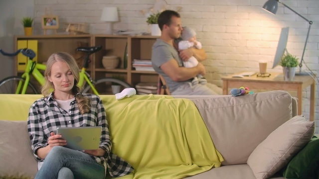 Beautiful mom sitting on couch with tablet while her husband and their little baby watching something on the computer 