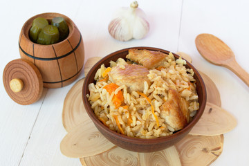 Pilaf with meat from a bowl on a wooden Board and white background, barrel pickles, garlic, wooden spoon