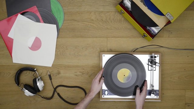 Man changing a vinyl record lifting it from the pickup and putting the album back in its sleeve. A pair of headphones and a stack of records on a wooden surface
