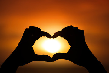 Silhouette of hand in heart shape with sun down in the middle 
