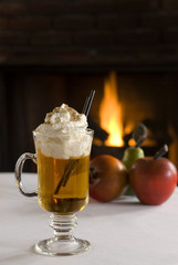 Hot apple cider drink topped with whipped cream and cinnamon. Fireplace and apples in the background.