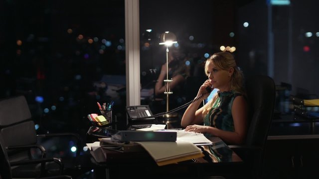 Beautiful business woman working overtime at night in executive office, answering phone call and talking on wired telephone. She feels stressed and tired. City lights in background