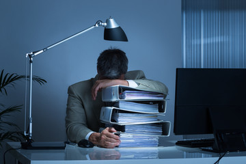 Businessman Leaning Head On Binders While Working Late