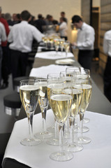 Eine Gruppe von gefüllten Sektgläsern oder Champagnergläsern im Vordergrund und eine Gruppe von Kellnern und Gästen im Hintergrund.