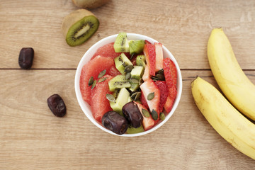 White bowl filled with fresh fruits, dried fruits, nuts, pumpkin seeds - grapefruit, bananas, strawberries, kiwi, dates. Wooden background.