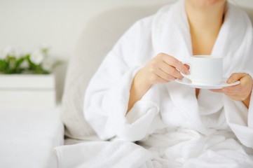 The woman in white bathrobe at the table drinking a hot drink.