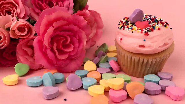 Valentine's Day scene with colorful candy hearts next to cupcake with bouquet of pink roses gets rained on candy sprinkles