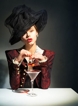 Woman in a vintage, black hat drinking a martini with a cherry