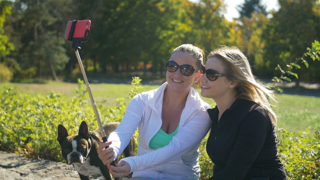 Two Pretty Young Women Taking Photos with Smart Phone on Selfie Stick. Blond and Brunette Taking Cell Phone Selfies on Sunny Day