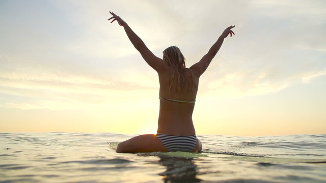 Young Happy Woman Celebrating at Sunset With Hands Up In Air on Surfboard. 