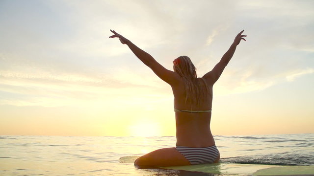 Young Happy Woman Celebrating at Sunset With Hands Up In Air on Surfboard. 
