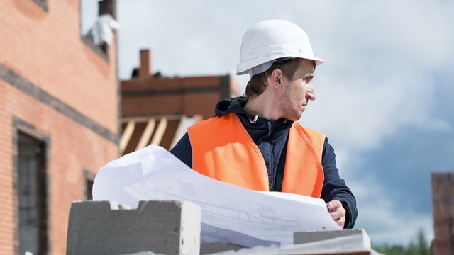 man engineer - builder at a construction site examines drawings or construction project. Looks thinking. The wind's blowing. Against the background of building object. Rain and clouds