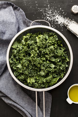 Fresh wet kale in a sieve, salt, olive oil and blue napkin on black chalkboard background captured...