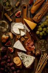 Different kinds of cheeses, wine, baguettes, fruits and snacks on rustic wooden table from above. French tasting party or feast scenery. Dark moody artistic style.