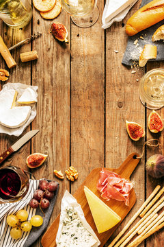 Different Kinds Of Cheeses, Wine, Baguettes, Fruits And Snacks On Rustic Wooden Table From Above. French Tasting Party Or Feast Scenery. Background Layout With Free Text Space.