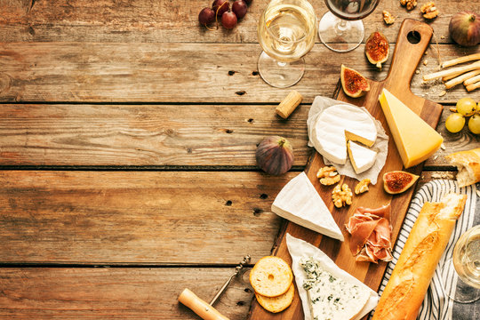 Different kinds of cheeses, wine, baguettes, fruits and snacks on rustic wooden table from above. French tasting party or feast scenery. Background layout with free text space.
