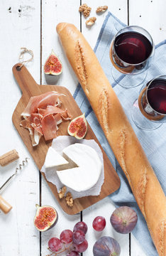 Camembert cheese, prosciutto (italian ham), baguette, two glasses of red wine, figs and grapes. White wooden table as background. Romantic french picnic scenery captured from above (top view).