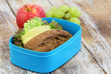 Healthy lunch box containing brown cheese sandwich, red apple and grapes on rustic wooden surface
