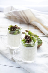 Vanilla panna cotta with kiwi and grated chocolate in glass, sliced kiwi on white wooden table