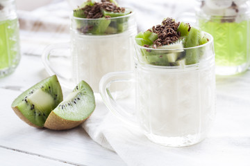 Vanilla panna cotta with kiwi and grated chocolate in glass, sliced kiwi on white wooden table