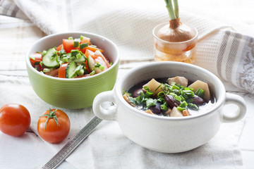 Vegetable soup with beans and chicken, fresh herbs in a clay pot, salad of tomatoes and cucumbers with greens in a green bowl on a white wooden table.