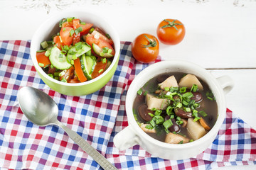 Vegetable soup with beans and chicken, fresh herbs in a clay pot, salad of tomatoes and cucumbers with greens in a green bowl on a white wooden table.