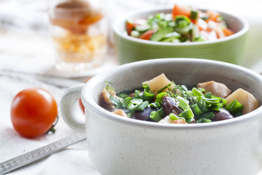 Vegetable soup with beans and chicken, fresh herbs in a clay pot, salad of tomatoes and cucumbers with greens in a green bowl on a white wooden table.