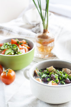 Vegetable soup with beans and chicken, fresh herbs in a clay pot, salad of tomatoes and cucumbers with greens in a green bowl on a white wooden table.
