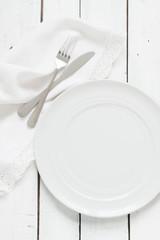 White table setting from above. Empty plate, cutlery, napkin on white planked wood table.