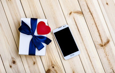 White Smart Phone With Blank Screen, White Gift Box With Blue Ribbon And Little Red Heart On Wooden Boards. Love concept. 