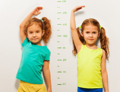Two girls show height on wall scale at home