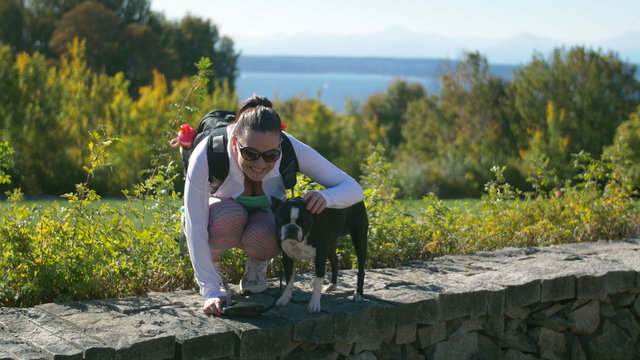 Woman Wearing Backpack with Dog Smiling For Camera in Front of Scenery. Brunette Young Lady Backpacking with Boston Terrier Posing for Photo with Ocean Background
