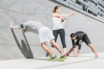 On a sunny day, a three friends in sportswear does stretching outdoors near a building